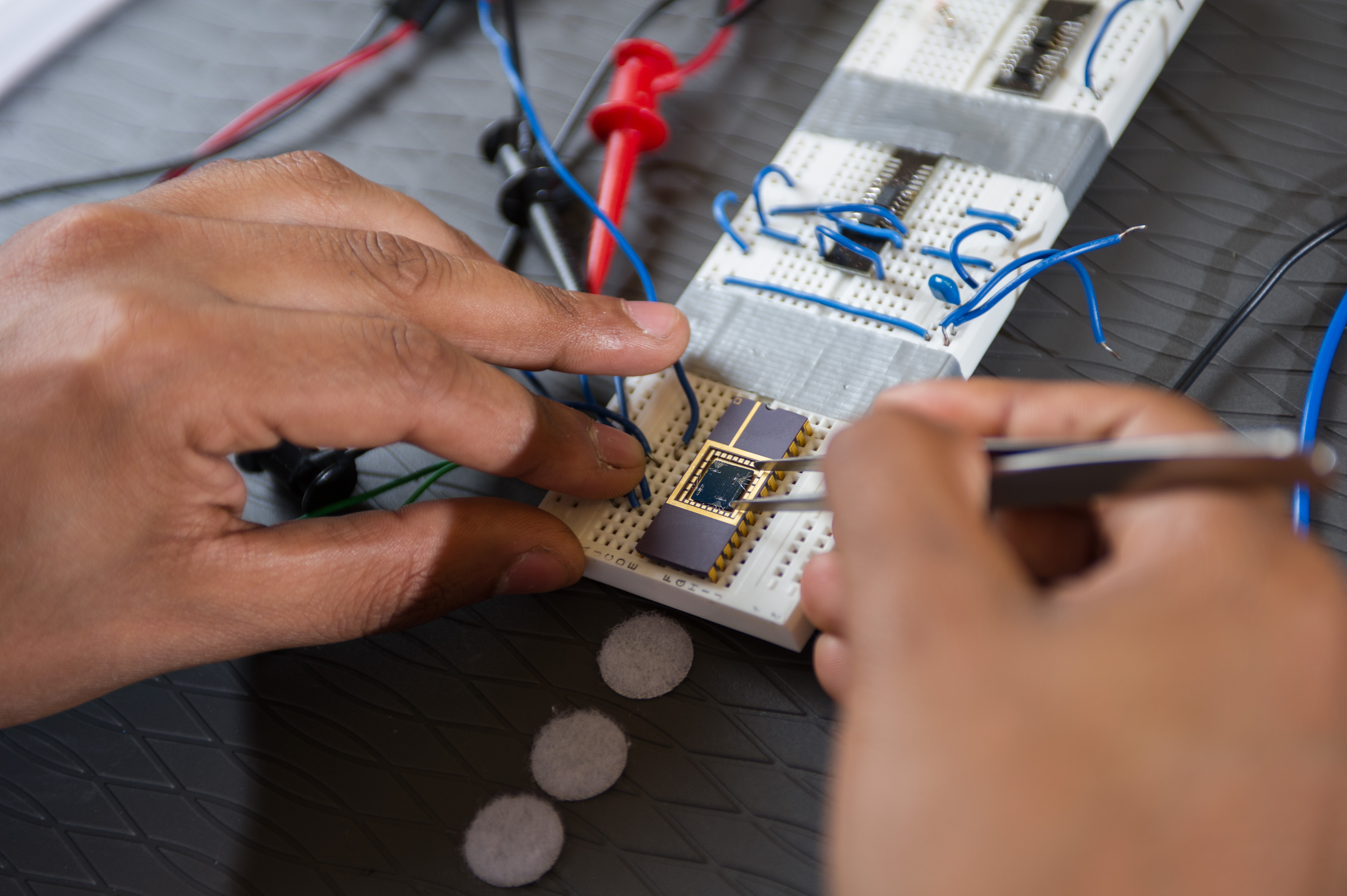 Close-up of hands assembling and testing a microelectronics circuit on breadboards with wires, probes, and a small semiconductor device in a laboratory setting.