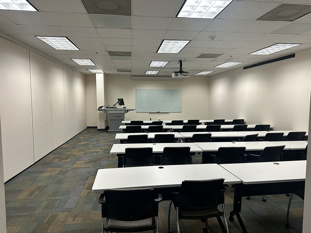 Empty classroom with tiered rows of tables and chairs, a lectern and whiteboard at the front, and fluorescent ceiling lights.