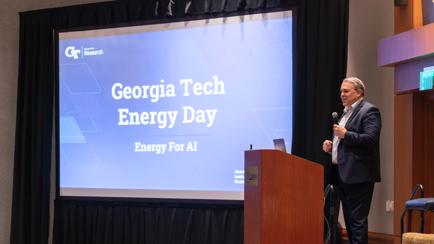 A man stands at a podium speaking in front of a large screen displaying “Georgia Tech Energy Day: Energy for AI.” The setting is a conference room with stage lighting and an audience out of frame.
