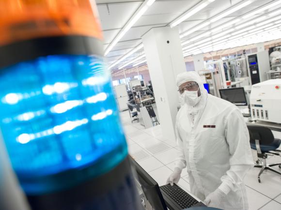A cleanroom technician in protective gear works at a computer workstation in a semiconductor lab, with a blue signal light in the foreground and lab equipment behind them.