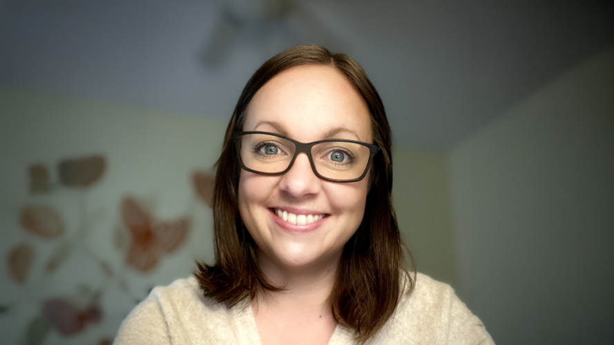 A woman wearing dark-framed glasses faces the camera against a softly blurred indoor background with muted blue-gray walls and abstract artwork.