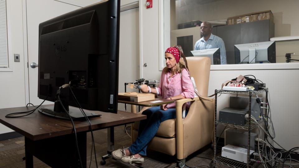A person seated in a beige chair using a computer setup with multiple cables and devices, facing a large monitor in a testing or research room, with another individual visible through a window in an adjacent control room.